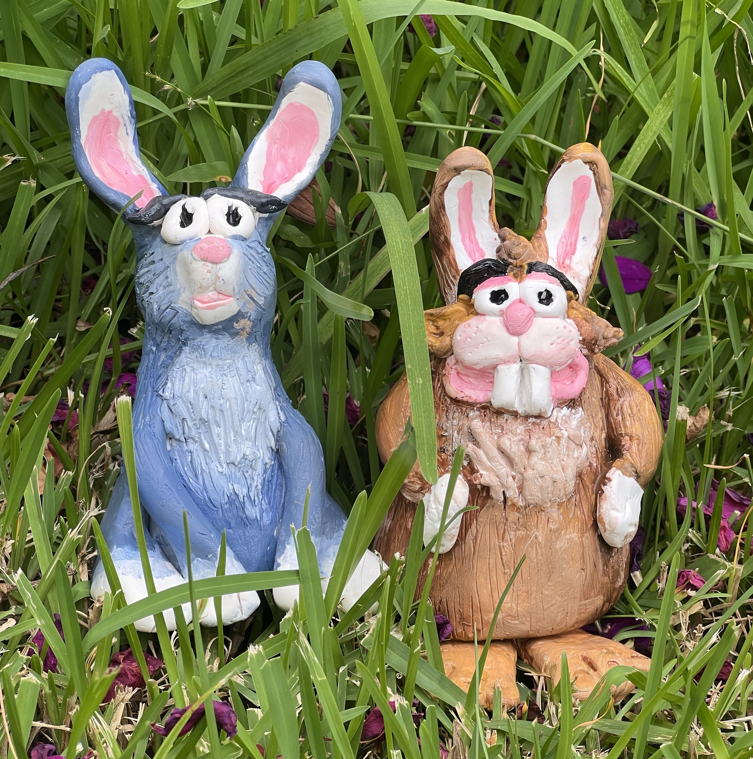 Two clay rabbits, one blue and one brown sitting in the green grass
