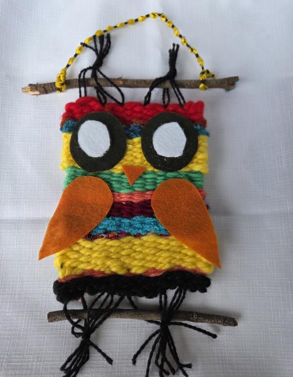 Woven owl, stripes of colour (yellow, blue, green, orange, red) hung between two sticks
