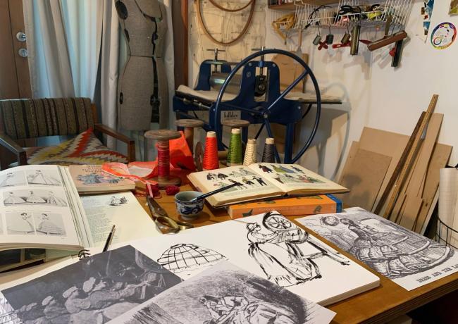 Photograph of a table covered in books, art sketches, and items like scissors and yarn.
