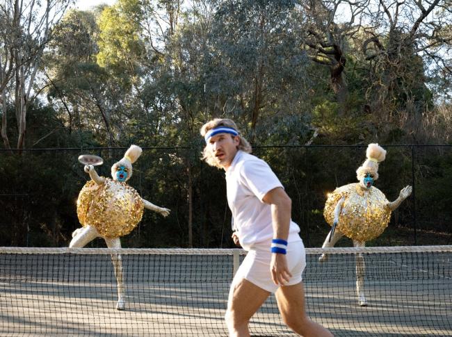 Three people playing tennis. Two of them are dressed in shiny round outfits and have their faces painted blue.